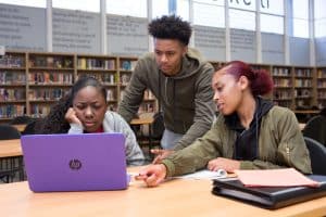 Three students collaborate on a computer in a library.