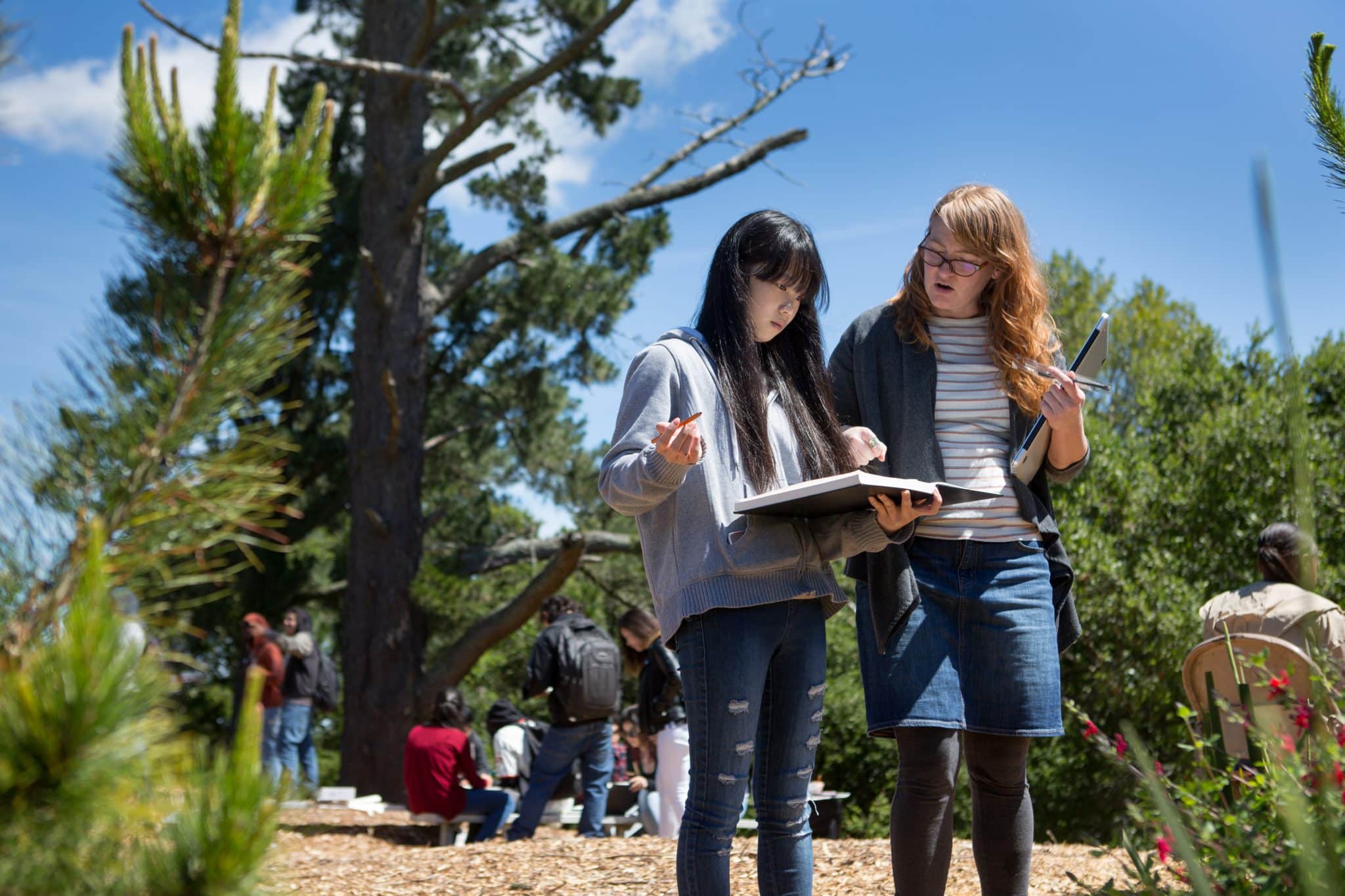 Teacher and Student Looking at Student's Artwork Outdoors with Other Students Doing Other Activities in the Background