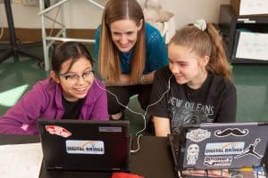 Female English Teacher With Two Female Students Sharing Earbuds