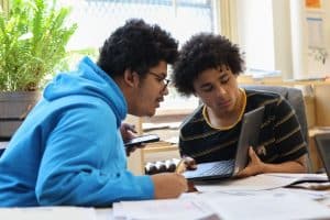 Two students working together, one using a phone and the other on a laptop, with papers on the desk and a fern in the background