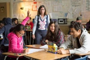 A student raises her hand while two others discuss at a desk, with a teacher observing in the background.