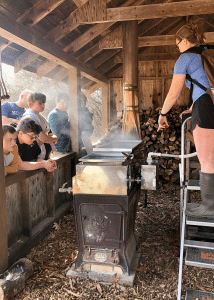 Students gather in a barn and look at a structure that looks to be a wood-burning stove.