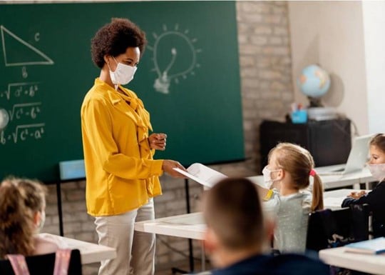 A teacher stands at the front of the classroom, handing a child at a desk a worksheet.