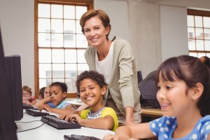 Cute pupil in computer class with teacher smiling at camera at t