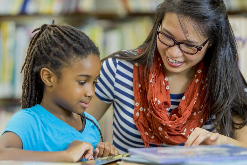 A teacher sits next to an elementary student, smiling and looking on while they read together.